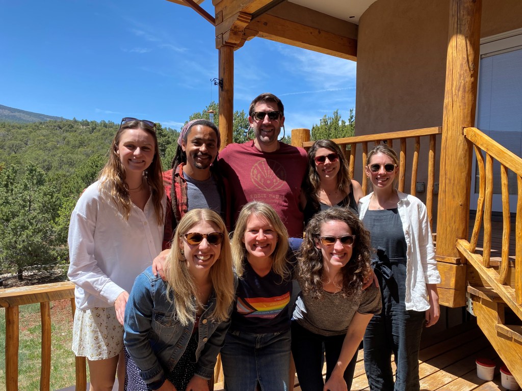 8 people on wood deck with mountain in background posing for picture.